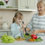 Little girl is cooking salad mixing vegetables in bowl and chatting to cheerful granny in kitchen at home. Modern lifestyle, meals preparation and family concept.